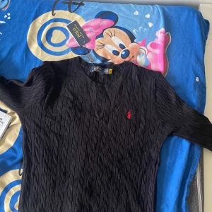 Svart kabelstickad tröja Polo Ralph Lauren - Svart kabelstickad tröja från Polo Ralph Lauren med klassisk rund halsringning och röd broderad logga på bröstet. Tröjan är i mjuk bomull och har långa ärmar. Perfekt för dig som gillar stilren och tidlös design.