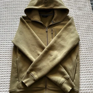 Olivgrön hoodie från Sail Racing S - Olivgrön hoodie från Sail Racing med broderad logga på bröstet. Tröjan har huva, lång ärm, bröstficka med dragkedja och mysigt fleecefoder på insidan. Perfekt för kyliga dagar och snygg till jeans eller joggers.