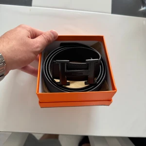 Svart skärp med H-spänne - Säljer ett stilrent svart bälte med ikoniskt H-spänne i metall. Bältet är tillverkat i exklusivt skinn och har en klassisk, rak form. Perfekt för dig som vill ha en lyxig accessoar med tydlig designer-vibe. 130 cm längd