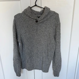 Grå stickad tröja från Dressmann - Mysig grå stickad tröja från Dressmann med snygg krage och knappdetalj vid halsen. Tröjan har lång ärm och ribbade muddar, perfekt för kyliga dagar. Enkel att matcha med jeans eller chinos för en avslappnad stil.