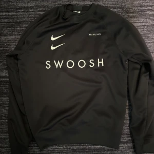 Svart Nike Swoosh sweatshirt - Svart sweatshirt från Nike med stora vita Swoosh-loggor fram och bak samt texten 'SWOOSH' på bröstet. Tröjan har rund hals, långa ärmar och är tillverkad i ett mjukt material som känns sportigt och bekvämt.
