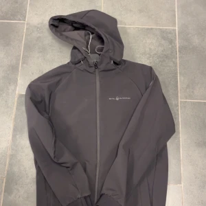 Svart vindjacka från Sail Racing - Svart vindjacka från Sail Racing med huva och hel dragkedja framtill. Jackan har en stilren design med logga på bröstet och elastiska muddar vid ärmslut och midja. Perfekt för blåsiga dagar och enkel att matcha med det mesta. Den har några defekter på armen längst ner men ingen som syns dirket när man har på sig den, storlek 170cm 