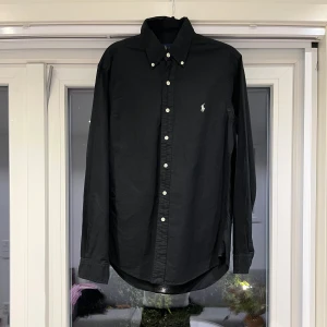 Svart skjorta från Ralph Lauren - Svart långärmad skjorta från Ralph Lauren i slim fit-modell. Klassisk button-down krage, vita knappar och broderad logga på bröstet. Skjortan är tillverkad i bomull och har en stilren, tidlös look som passar perfekt till jeans eller chinos.