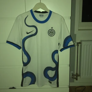 Inter Milan vit 21/22 bortatröja  Nike XS - Inter Milans officiella bortatröja från Nike i vitt med blå och svarta detaljer. Tröjan har ett unikt ormmönster, klubbmärke på bröstet och Lenovo-tryck på ryggen. Tillverkad i Dri-FIT-material som andas och har blå ribbad krage och ärmslut.