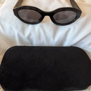 Svarta ovala solglasögon från Gucci - Snygga svarta solglasögon från Gucci med oval form och breda skalmar. Guldig Gucci-logga på sidan och mörka glas. Tillverkade i Italien och levereras med svart fodral. Perfekt accessoar för en trendig look.
