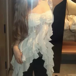 Vit spetsklänning med volanger M - Romantisk vit klänning med spets och volanger längs sidorna. Klänningen har smala axelband med volangdetaljer och en söt blomapplikation framtill. Materialet är lätt och luftigt, perfekt för sommarens varma dagar. Den har en feminin och drömmig look med omlottkänsla. Korea brand