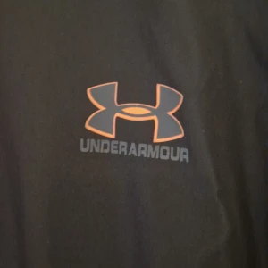 Svart träningsjacka från Under Armour - Svart träningsjacka från Under Armour med orange dragkedja och detaljer. Jackan har logga på bröstet och ärmen, samt gråa och orangea inslag på ärmen. Perfekt för dig som gillar sportig stil och vill sticka ut med snygga kontraster.