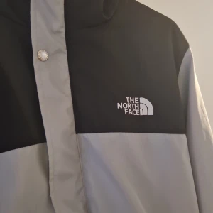 Svart och grå vindjacka The North Face - Snygg tvåfärgad vindjacka från The North Face i svart och grått. Jackan har huva med dragsko, dragkedja och tryckknappar framtill samt två fickor med dragkedja. Perfekt för blåsiga dagar och streetwear-stil. Funkar för dom som har strl M oxå