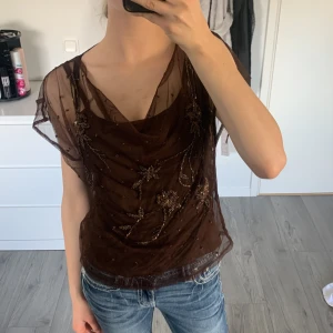 Vintage mesh blus med broderade blommor - Säljer en vintage brun transparent meshblus med broderade blommor och pärldetaljer. Blusen har korta ärmar och ett lätt draperat fall framtill. Storlek M men passar mig helt okej som vanligtvis har XS/S. Nypris är förmodligen mellan 400 och 700 kr. Hör av dig vid frågor💕