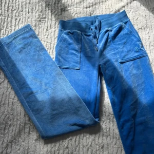 Blå mjukisbyxor från Juicy Couture - Blå mjukisbyxor från Juicy Couture med bred resår och snörning i midjan. Byxorna har raka ben, stora fickor framtill och broderad JC-logga på ena fickan. Tillverkade i mjukt och skönt material som känns lyxigt mot huden.