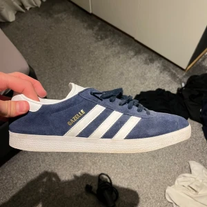Adidas Gazelle blå sneakers - Säljer ett par klassiska Adidas Gazelle sneakers i marinblå mocka med vita detaljer och guldtext på sidan. Skorna har de ikoniska tre ränderna, vit platt sula och rund tå. Perfekt för dig som gillar retrostil och snygga färgkombinationer.