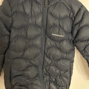 Svart dunjacka Peak Performance - Snygg mörkblå dunjacka från Peak Performance med vågformat quiltat mönster. Jackan har dragkedja framtill, två fickor med dragkedja och diskret logga på bröstet. Perfekt för kalla dagar och riktigt skön att bära. Sitter som en S
