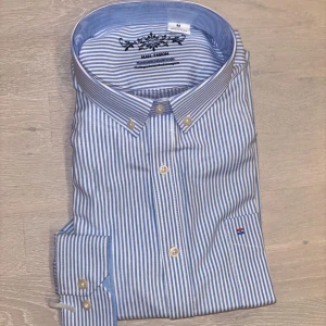 Randig skjorta - Klassisk skjorta i blåvitt randigt mönster. Skjortan har button down-krage, långa ärmar och liten broderad logga på bröstet. Tillverkad i mjuk bomull och har snygga vita knappar på både framsidan och manschetterna. Aldrig använd och kragens skydd sitter kvar
