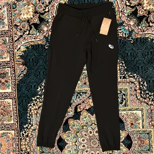 Träningsbyxor från Asics - Svarta byxor från Asics med elastisk midja och snörning. Byxorna har smal passform, muddar vid bensluten och en vit logga på låret. Perfekta för sommar eller träning