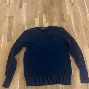 Mörkblå kabelstickad tröja Polo Ralph Lauren - Kabelstickad mörkblå tröja från Polo Ralph Lauren i 100% silke. Klassisk rund halsringning, ribbade muddar och röd broderad logga på bröstet. Perfekt för dig  som gillar stilren och tidlös design.  
