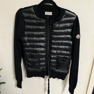 Svart cardigan jacka från Moncler - Svart cardigan jacka från Moncler med quiltad front i glansigt material och stickade ärmar. Jackan har två fickor framtill med knapp, dragkedja och Moncler-logga på ärmen. Perfekt för dig som vill ha en stilren och sportig look.