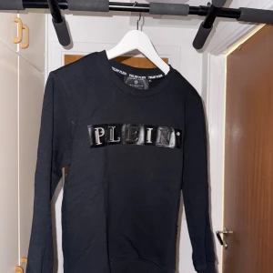 Philipp Plein tröja - Svart sweatshirt från Philipp Plein med glansig svart patch och silverfärgad text 'PLEIN' framtill. Klassisk rund halsringning och lång ärm. Snygg detalj med metallmärke i nacken. Perfekt för dig som gillar streetwear med exklusiv känsla. Säljer då den är för liten på mig, kontakta mig för frågor m.m!!