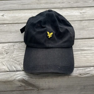 Svart keps från Lyle & Scott - Svart keps från Lyle & Scott med klassisk böjd skärm och justerbart spänne bak. Framtill finns en gul broderad örnlogga. Kepsen är tillverkad i bomull och har ventilerande hål för extra komfort. Perfekt accessoar för en avslappnad stil.