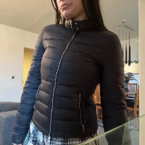 Zara Svart dunjacka med dragkedja - Snygg svart Zara  dunjacka med smal passform och horisontella sömmar. Jackan har hög krage med tryckknappar och dragkedja framtill samt dragkedjeförsedda fickor. Perfekt för kyliga dagar och enkel att matcha med olika outfits.