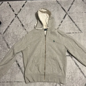 Grå hoodie från Polo Ralph Lauren - Snygg ljusgrå hoodie från Polo Ralph Lauren med dragkedja och klassisk grön logga på bröstet. Tröjan har huva med vit insida och snörning samt två fickor framtill. Perfekt för chill dagar och enkel att matcha med jeans eller joggers.