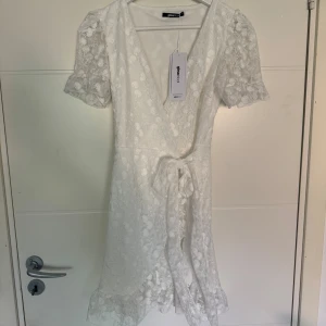 Vit omlottklänning i spets  - Supersöt vit kortklänning från Gina Tricot med blommigt spetsmönster, puffiga kortärmar och v-ringning. Omlottmodell med knyt i midjan och volangdetalj längst ner. Perfekt för studenten! Helt ny med lappen kvar.