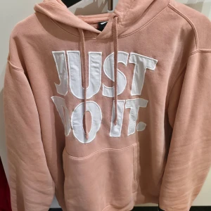 Nike hoodie i ljusrosa med tryck - Nike hoodie i ljusrosa med stort vitt 'JUST DO IT.'-tryck på bröstet. Klassisk känguruficka, huva med snörning och mjukt material i 80% bomull och 20% polyester. Perfekt för chill dagar och streetstyle.
