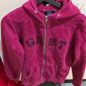 Vinröd hoodie från GANT med dragkedja - Säljer en vinröd hoodie från GANT med broderad logga framtill. Tröjan har huva, fickor och hel dragkedja. Tillverkad i mjuk bomull och polyesterblandning, perfekt för kyliga dagar. Snygg och bekväm passform med klassisk collegekänsla.