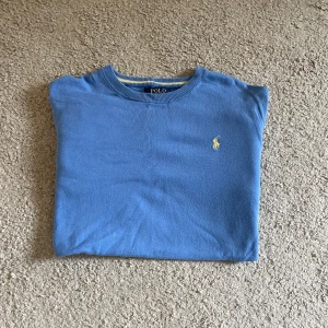 Blå tröja från Ralph Lauren - Snygg blå stickad tröja från Polo Ralph Lauren med klassisk gul broderad logga på bröstet. Tröjan har rund halsringning, ribbade muddar och är långärmad. Perfekt för dig som gillar stilrena och tidlösa plagg.