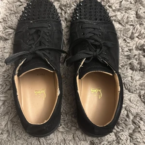 Christian Louboutin svarta sneakers med nitar - Svarta sneakers från Christian Louboutin i mocka med ikoniska röda sulor och svarta nitar på tån. Skorna har snörning och rund tå, samt coola detaljer som ger en edgy vibe. Perfekta för dig som vill sticka ut med lyxig streetstyle.