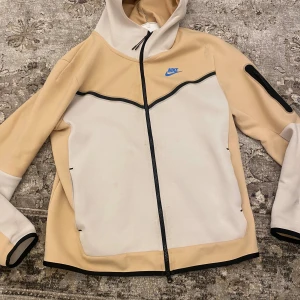 Beige och vit Nike vindjacka med huva - Nike vindjacka i beige och vitt med svart dragkedja och blå Nike-logga på bröstet. Jackan har huva, lång ärm med ficka på ena ärmen och sportig design. Perfekt för dig som gillar streetwear och vill ha en clean look.