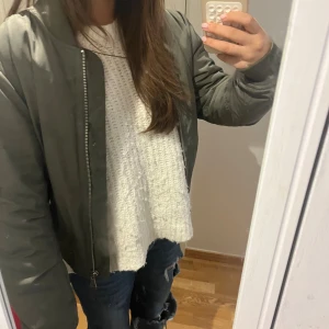 Grön Bomberjacka  - Säljer en khakigrön bomberjacka med klassisk passform och ribbade muddar. Jackan har dragkedja framtill och en extra ficka med dragkedja på ärmen. Materialet är slätt och lätt glansigt, perfekt för höst och vår.