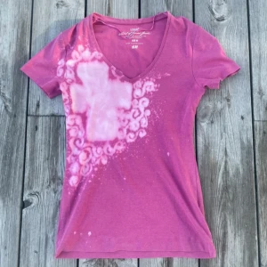 Rosa t-shirt med kors och vingar H&M - Cool rosa t-shirt med v-ringning och korta ärmar. Framsidan har ett blekt kors och swirlmönster, baksidan har blekta änglavingar. Skön passform och tillverkad i mjuk bomull. Perfekt för dig som gillar unika detaljer och statement-plagg. köpt secondhand och sedan omgjord. lite nopprig men tycker de passar med designen