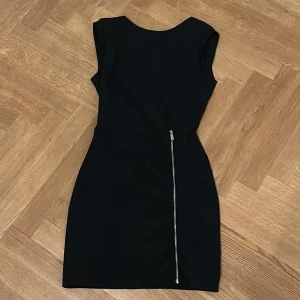 Svart kort klänning med dragkedja - En svart, figurnära kort klänning med ärmlös design och en snygg dragkedja som löper diagonalt längs baksidan. Perfekt för dig som gillar stilrena och moderna plagg med en edgy detalj.
