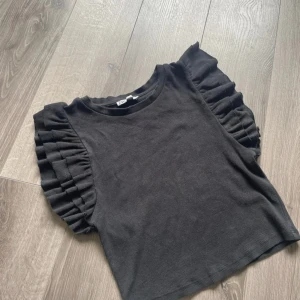 Svart volangtopp från Zara - Svart topp från Zara med korta ärmar och snygga volangdetaljer på axlarna. Toppen är ribbad och har en croppad passform som ger en trendig look. Perfekt att styla med jeans eller kjol för en cool outfit.