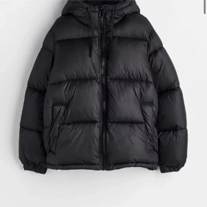 h&m jacka - Säljer en svart pufferjacka med huva och dragkedja framtill. Jackan är vadderad och har en oversized look med långa ärmar och två sidofickor. Perfekt för kalla dagar när du vill hålla dig varm och ändå se snygg ut.