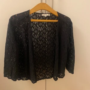 Svart spetskofta  - Svart kofta i spets från Flash Woman med blommigt mönster. Koftan har trekvartsärmar och är öppen framtill, perfekt att slänga över en topp för en snygg layering. 