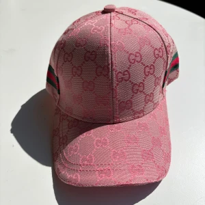 Rosa keps från Gucci med GG-mönster - Säljer en rosa keps från Gucci med ikoniskt GG-monogram över hela kepsen. Kepsen har gröna och röda ränder på sidorna och justerbar rem bak. Tillverkad i bomull och polyester, perfekt för dig som vill sticka ut med en lyxig accessoar.