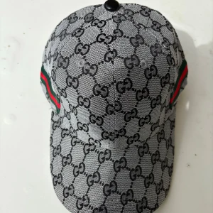 Gucci keps med GG-mönster - Snygg keps från Gucci i grått och svart med klassiskt GG-monogram över hela kepsen. Har gröna och röda ränder på sidorna och justerbar rem i svart skinn baktill. Tillverkad i bomull och skinn, perfekt för dig som vill sticka ut med en lyxig accessoar.