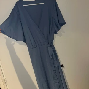 Omlottklänning med knytband - Snygg blå långklänning med omlottmodell och v-ringning. Klänningen har vida, korta ärmar och ett knytband i midjan som ger en fin siluett. Perfekt för dig som gillar en enkel men elegant stil.