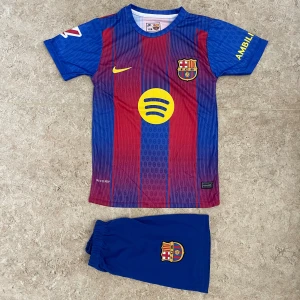 FC Barcelona Lamine Yamal fotbollströja - Säljer en FC Barcelona matchtröja med Lamine Yamal och nummer 10 på ryggen. Tröjan är kortärmad, har klassiska blå och röda ränder, gula detaljer och klubbmärke på bröstet. Materialet är lätt och andas, perfekt för fotboll. Shorts med klubbmärke ingår.