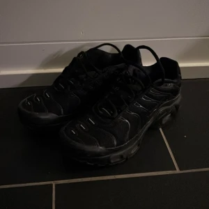 Nike Air Max Plus TN helsvarta sneakers - Säljer ett par helsvarta Nike Air Max Plus TN sneakers med snygga vågformade detaljer och Tn-logga bak. Skorna har snörning, platt sula och är tillverkade i syntet och mesh för en sportig look. Perfekta för dig som gillar streetstyle och vill ha något som sticker ut. 