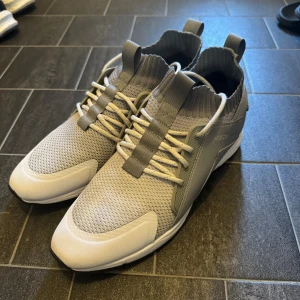 Grå sneakers från Hugo med mesh - Snygga grå sneakers från Hugo med vit sula och stickad, ribbad kant runt ankeln. Ovandelen är i mesh och syntet, med tydlig logga bak och på sidan. Skorna har snörning och dragflikar både fram och bak för enkel påtagning. Perfekta för en clean och modern look.