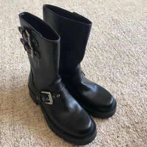 Zara Svarta boots med spännen i skinn - Tuffa svarta boots i skinn med grov sula och tre silvriga spännen på varje sko. Modellen har rund tå och platt sula, perfekt för dig som gillar en edgy stil. Passar till både jeans och kjol för en cool look.
