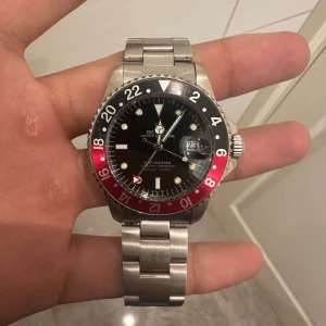 Klocka GMT-Master svart/röd stål - Klockan är en  GMT-Master med klassisk svart urtavla och röd/svart vridring. Klockan har en robust länk i stål och tydlig datumvisning med förstoringsglas. Ikonisk design med tydliga index och extra GMT-visare. Perfekt för dig som gillar stilren och sportig look. Den fungerar bra. Det är ett automatisk urverk. I bra skick. Om ni har andra frågor de ba ställa.  kan hämtas upp i Stockholm/årsta om man vill 