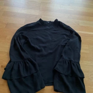 Svart blus med volangärmar - Svart blus med långa ärmar med dubbla volanger. Blusen har en knapp i nacken och är i mjukt, lätt material. 