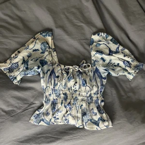 Blommig croppad blus med puffärm - Säljer en ljusblå och vit blommig blus med croppad passform och puffiga korta ärmar. Toppen har smockad midja, volangkant och en söt snörning framtill vid halsen. Perfekt för dig som gillar romantisk och trendig stil.