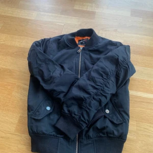 Svart bomberjacka - Svart bomberjacka. Jackan har orange insida, ribbade muddar och dragkedja framtill. Två fickor med tryckknappar. 