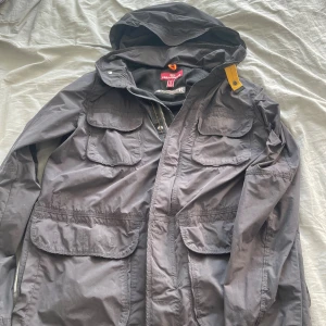 Svart vindjacka från Parajumpers - Säljer en svart vindjacka från Parajumpers med huva och flera stora fickor framtill. Jackan har en gul detalj vid axeln och stängs med dragkedja och tryckknappar. Materialet är lätt och perfekt för blåsiga dagar. Klassisk streetstil med snygga detaljer.