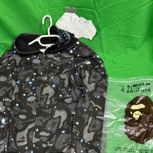 A Bathing Ape camo zip hoodie - Säljer en fet svart och grå camo zip hoodie från BAPE med blå detaljer och ikonisk hajdesign på huvan. Jackan har dragkedja hela vägen, huva och ett unikt camomönster med inslag av blått och vitt. Perfekt för dig som vill sticka ut med streetwear.Liknande modell som originalet!