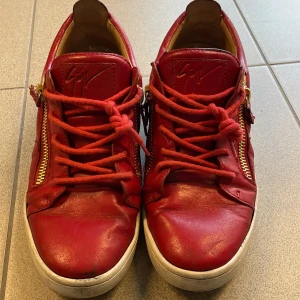 Röda sneakers från Giuseppe Zanotti - Säljer ett par röda sneakers från Giuseppe Zanotti med coola guldfärgade dragkedjor på sidorna och matchande röda snören. Skorna är tillverkade i skinn och har en vit platt sula. Ikonisk logga på plösen och snygg rund tå. Perfekt för dig som gillar statement pieces. Perfekt för en unik stil och varma perfekta skor till vintern